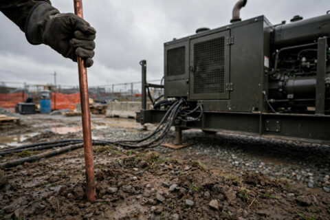 Copper ground rod being driven into soil next to an industrial diesel generator on a construction site — documentary industrial photography